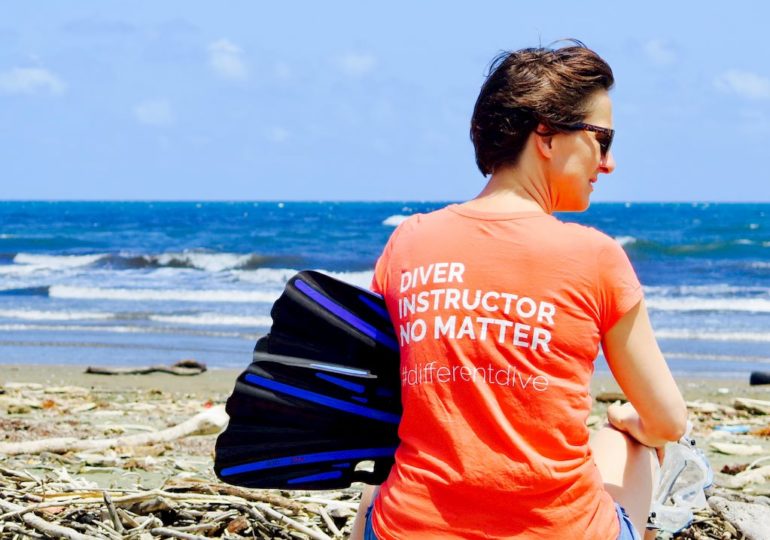 Hélène assise sur la plage avec ses palmes et son masque. Elle arbore un t-shirt avec écrit dessus "divers, instructeur, no matter #differentdive"