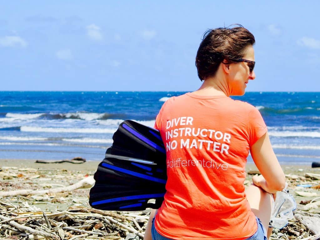 Hélène assise sur la plage avec ses palmes et son masque. Elle arbore un t-shirt avec écrit dessus "divers, instructeur, no matter #differentdive"